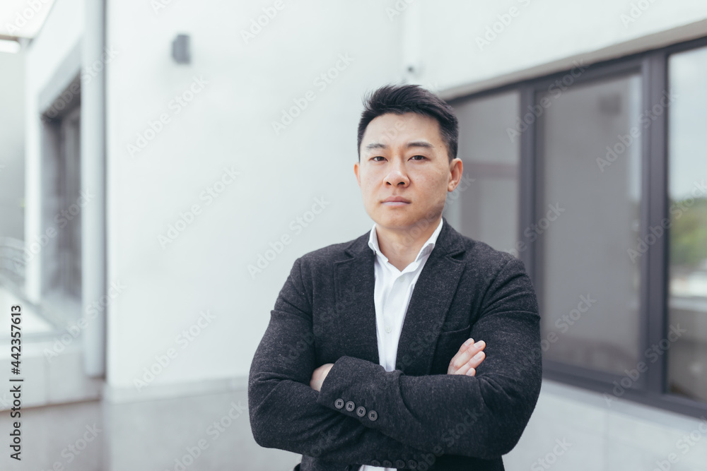 Young Asian businessman looks at the camera in a business suit, arms folded crosswise near the office center Young Asian businessman looks at the camera in a business suit, arms folded crosswise near the office center