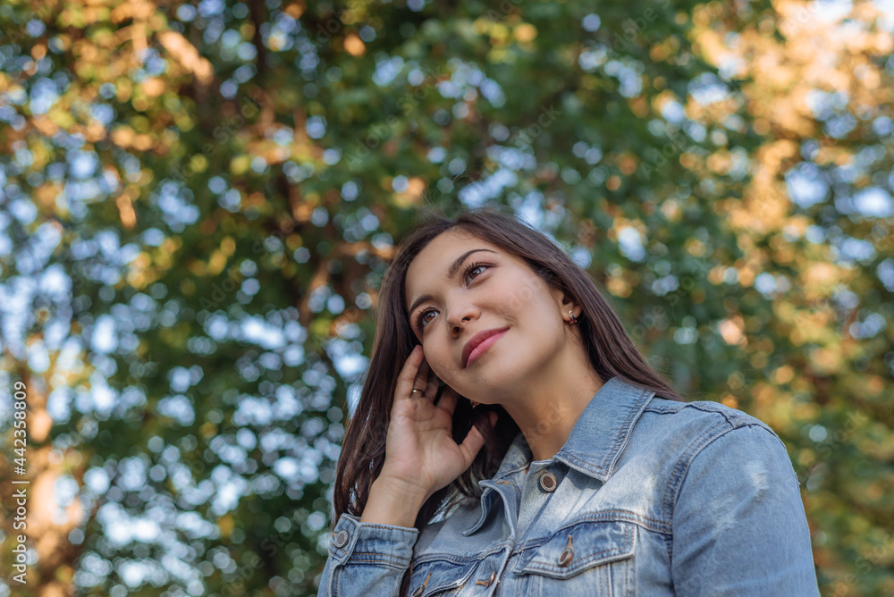 portrait of a beautiful young Asian woman with dark hair in a park. a pretty Asian woman in a denim jacket against the background of green trees smiles portrait of a beautiful young Asian woman with dark hair in a park. a pretty Asian woman in a denim jacket against the background of green trees smiles