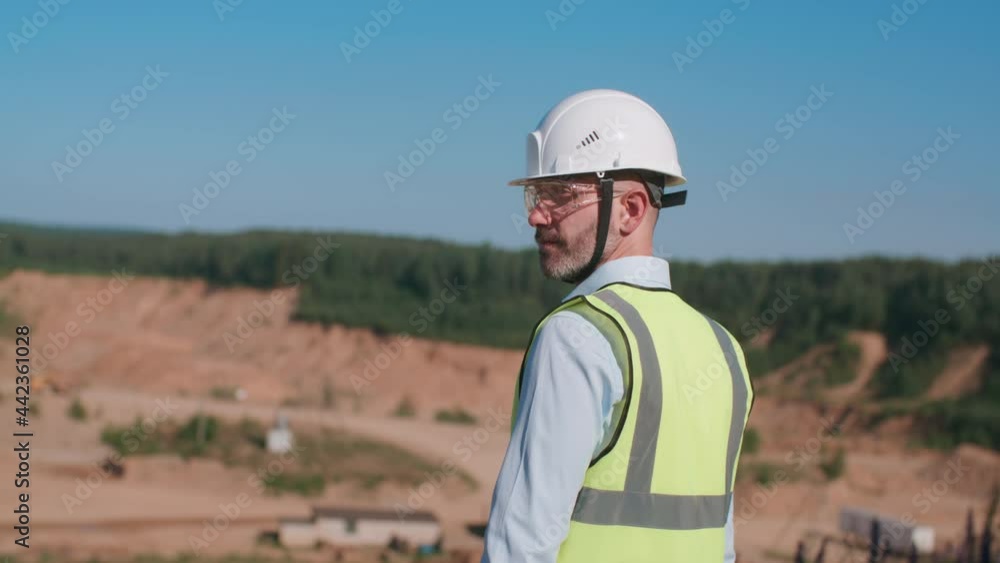 Handheld following handsome sand quarry worker engineer, male caucasian foreman in safety wear and hardhat walks along construction site. Bearded industrial worker builder, mining project specialist Handheld following handsome sand quarry worker engineer, male caucasian foreman in safety wear and hardhat walks along construction site. Bearded industrial worker builder, mining project specialist
