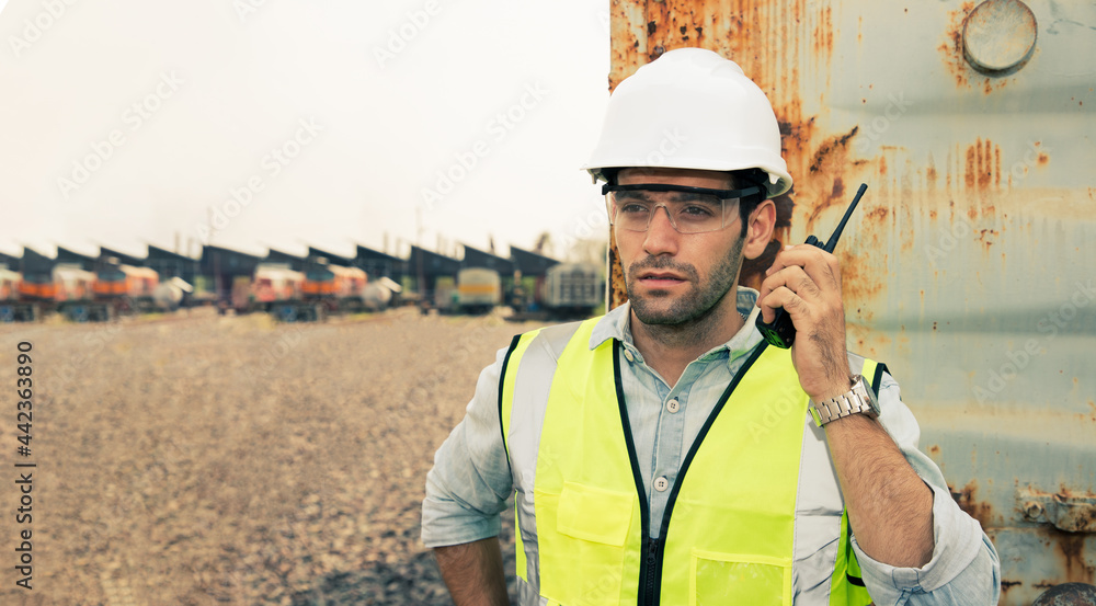 Portrait of handsome engineering using walkie talkie for control labor with wear hardhat in front of train garage.  Back view of contractor on background of outdoor old train garage.