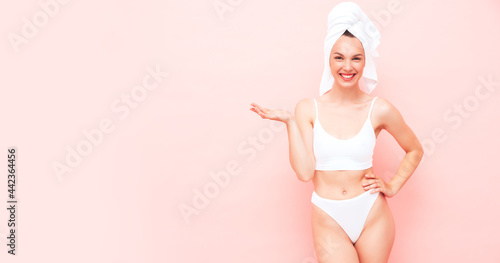Young beautiful smiling woman in white lingerie. Sexy carefree model in underwear and towel on head posing pink wall in studio. Positive and happy female enjoying morning. Shows something on her hand