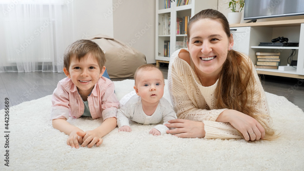 Happy smiling mother with baby and older son lying on soft carpet at living room. Parenting, children happiness and family relationship