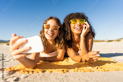 Front portrait of two cute girls with sunglasses in summer vacation using smartphone taking a selfie lying on sea sand of a tropical beach. Multiracial women travellers having fun with technology
