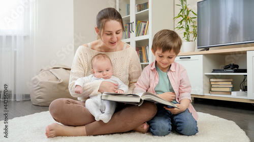 Fényképezés Smiling mother sitting on floor and reading story book to her older and younger sons