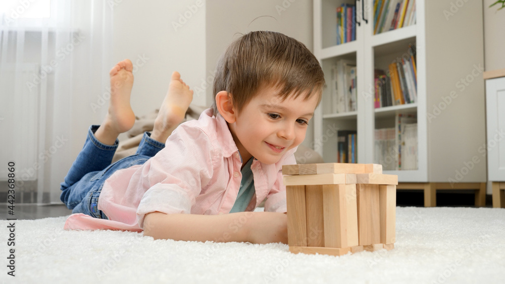Smiling boy lying on floor and building house or tower from wooden blocks. Concept of child education and games at home Smiling boy lying on floor and building house or tower from wooden blocks. Concept of child education and games at home