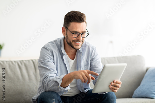 Free time concept. Happy man using digital tablet, sitting on couch in living room, copy space