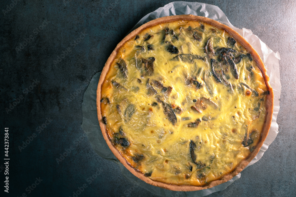 Top view of a freshly baked yellow quiche with mushrooms still on a baking paper, sitting on a dark kitchen table surface. Top view of a freshly baked yellow quiche with mushrooms still on a baking paper, sitting on a dark kitchen table surface.