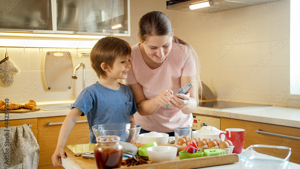 Happy little boy with young mother looking for new recipe to cook on smartphone. Children cooking with parents, little chef, family having time together, domestic kitchen. Happy little boy with young mother looking for new recipe to cook on smartphone. Children cooking with parents, little chef, family having time together, domestic kitchen.