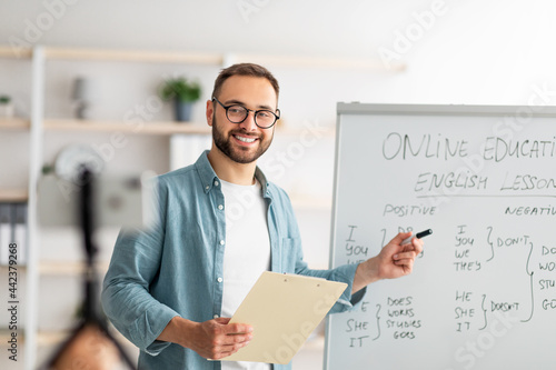 Portrait of happy young male teacher standing near blackboard with English grammar rules, conducting internet lesson