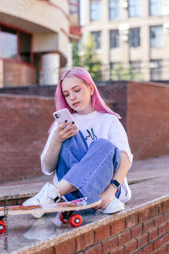 A pink-haired hipster teenage girl in a white T-shirt,jeans and with a skateboard is sitting on a city street on a summer day and using a smartphone.Generation Z style,social network,hobby.