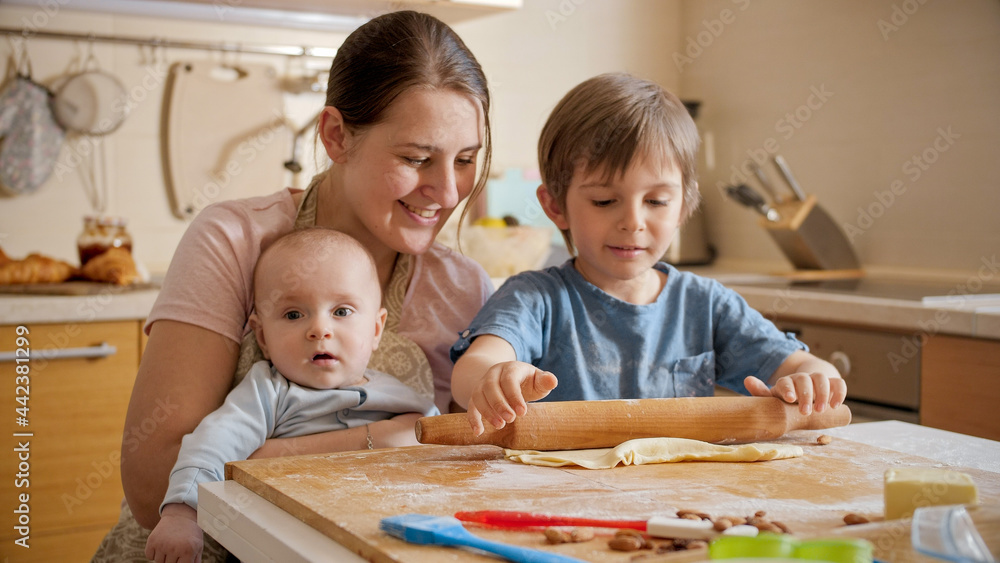Young mother holding baby helping older son baking pizza at home. Children cooking with parents, little chef, family having time together, domestic kitchen. Young mother holding baby helping older son baking pizza at home. Children cooking with parents, little chef, family having time together, domestic kitchen.