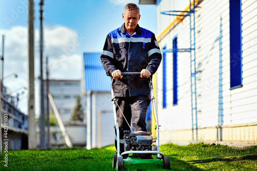 Fotografie A pensioner in working clothes works with a lawn mower on the territory of the production base on a sunny day