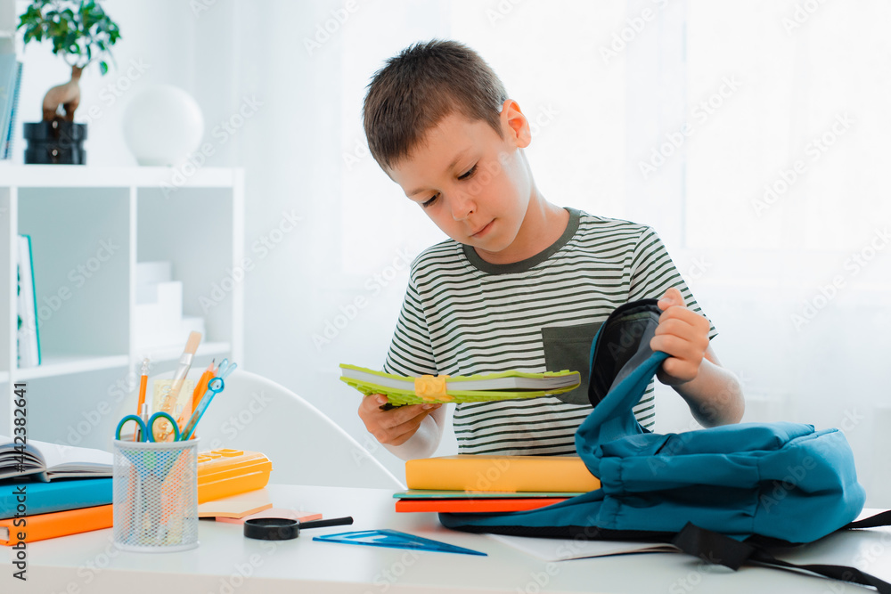 schoolboy putting school stationery into backpack at table indoors at home white room. schoolboy putting school stationery into backpack at table indoors at home white room.