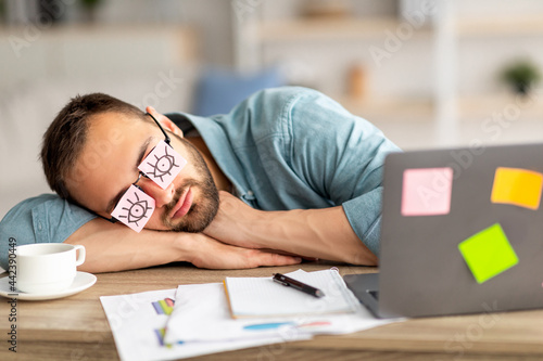 Lazy unproductive young guy wearing funny sticky notes with open eyes on his glasses, sleeping at workplace photo