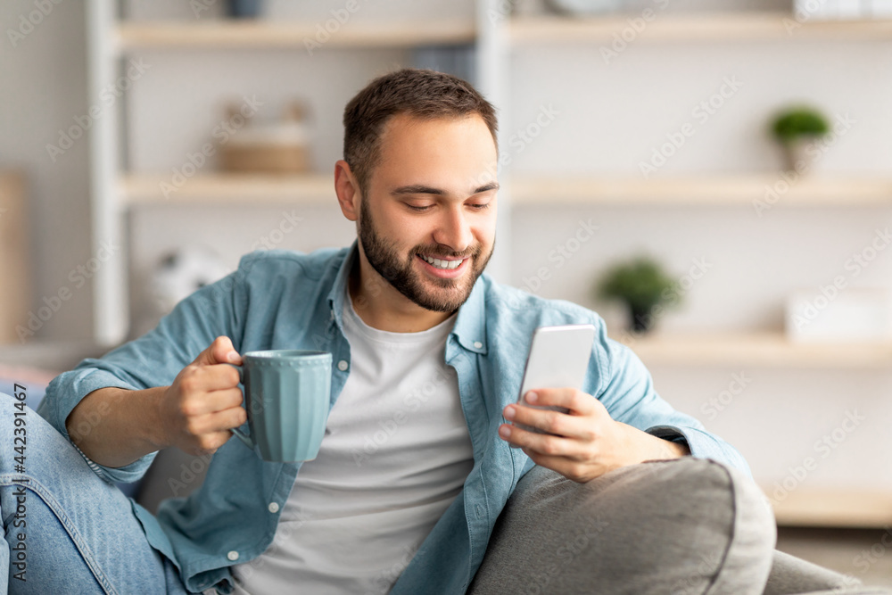Positive young guy holding smartphone and coffee indoors, chatting with friends online, sending email, browsing internet Positive young guy holding smartphone and coffee indoors, chatting with friends online, sending email, browsing internet