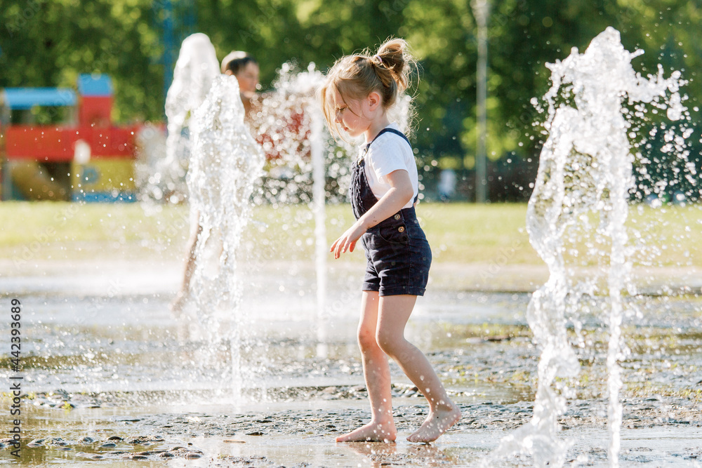 Cute cheerful girl playing in fountain. Kid in denim overall having fun in summer park