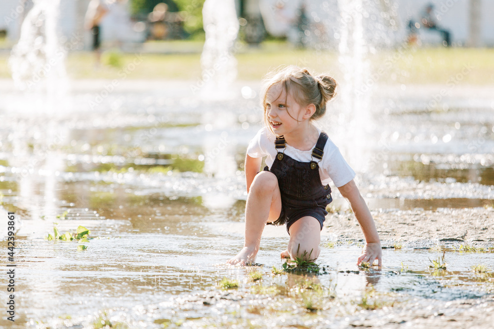 Cute cheerful girl playing in fountain. Kid in denim overall having fun in summer park