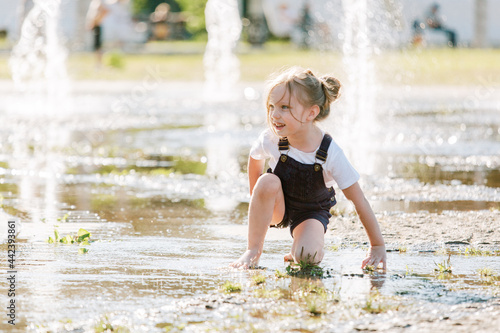 Obraz na płótnie Cute cheerful girl playing in fountain. Kid in denim overall having fun in summer park w salonie