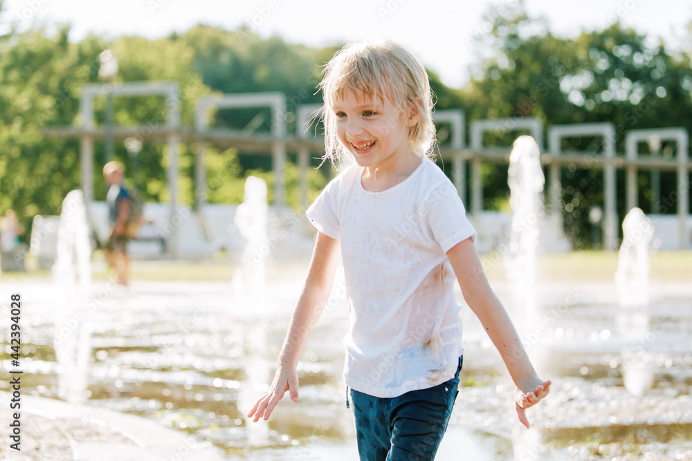Cute cheerful girl playing in fountain. Kid in white shirt having fun in summer park