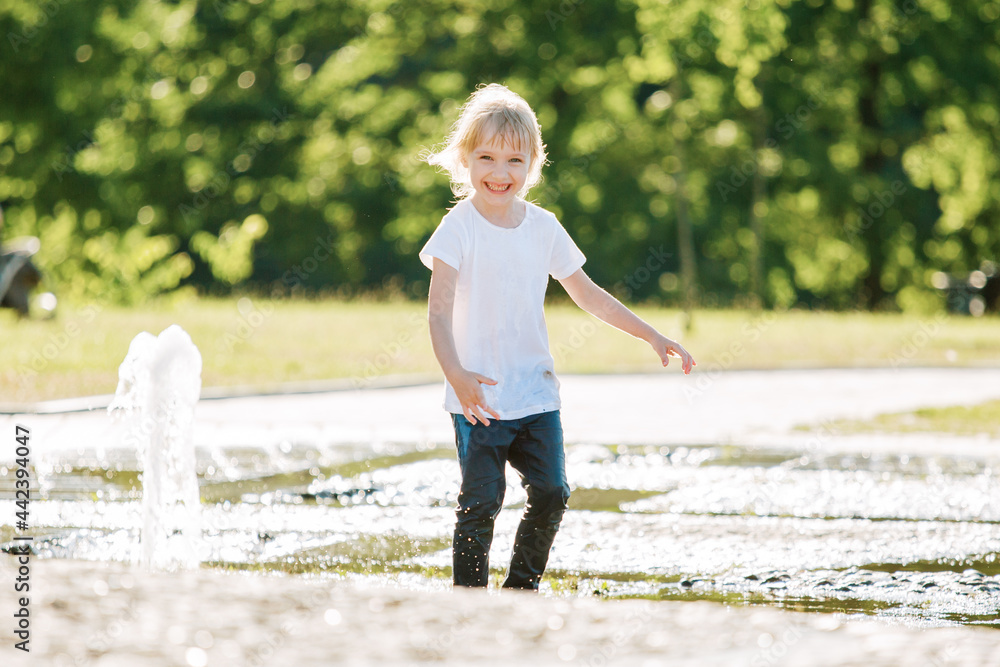 Cute cheerful girl playing in fountain. Kid in white shirt having fun in summer park