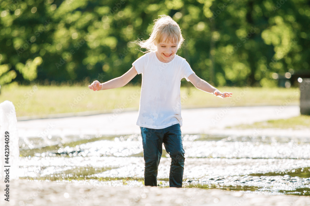 Cute cheerful girl playing in fountain. Kid in white shirt having fun in summer park