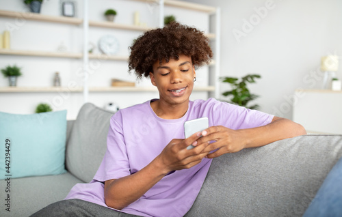 Cool black teenager with mobile phone studying online, chatting with friend, posting in social media from home