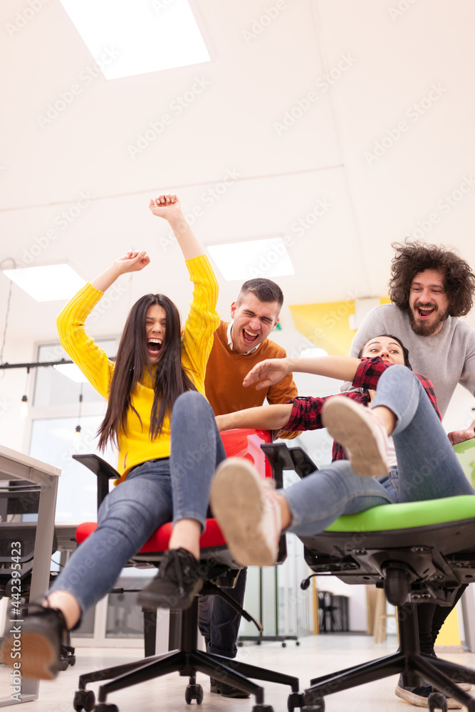 Colleagues having fun on work at the office. Girls pushing boys on chairs with wheels. Multiethnic group of successful start up company. Colleagues having fun on work at the office. Girls pushing boys on chairs with wheels. Multiethnic group of successful start up company.