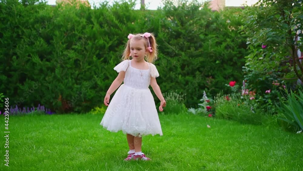 A beautiful little girl in a white dress stands on the green lawn in the garden. A beautiful little girl in a white dress stands on the green lawn in the garden.