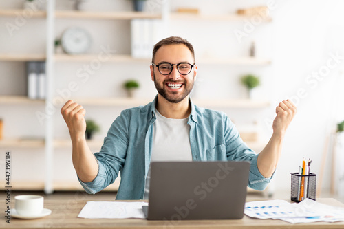 Young guy making YES gesture in front of laptop, reaching success, making profitable online deal at home office