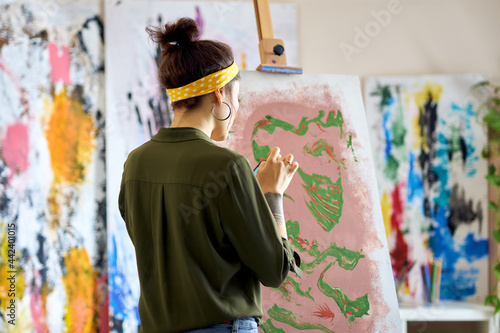 Back view of young female artist creating abstract painting, standing in living room at home