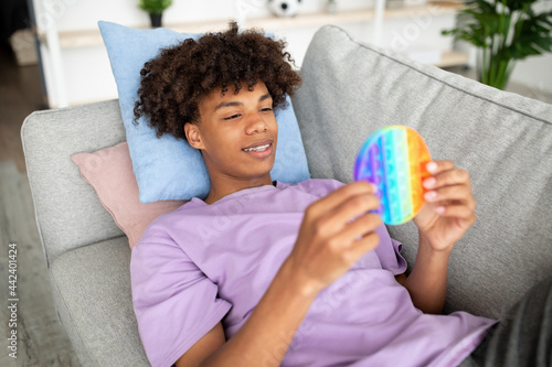 Cool African American teen lying on comfy sofa, playing with trendy POP IT toy at home photo