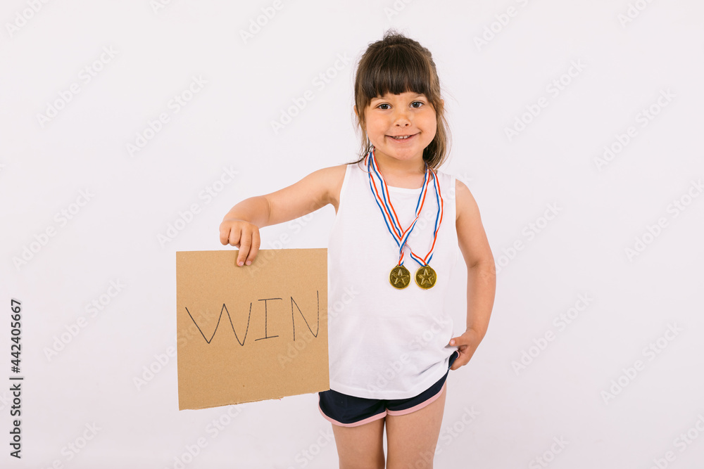 Small dark-haired girl with sports champion medals, holding a sign that reads: 'win'. Sport and victory concept Small dark-haired girl with sports champion medals, holding a sign that reads: 'win'. Sport and victory concept