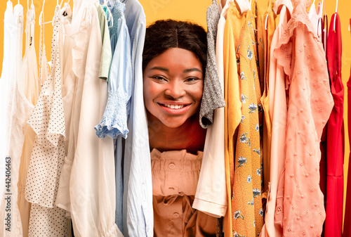 Young black woman peeking out between clothes hanging in rail, smiling at camera, satisfied with shopping choice