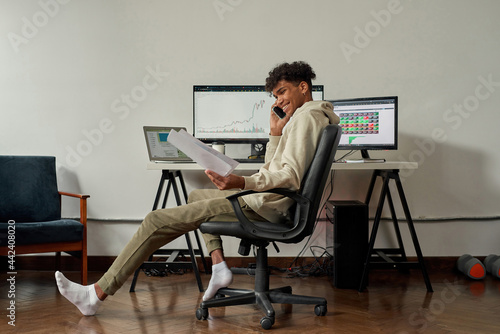 Full length shot of relaxed young male trader holding papers with statistics while talking on the phone, sitting at desk, trading from home