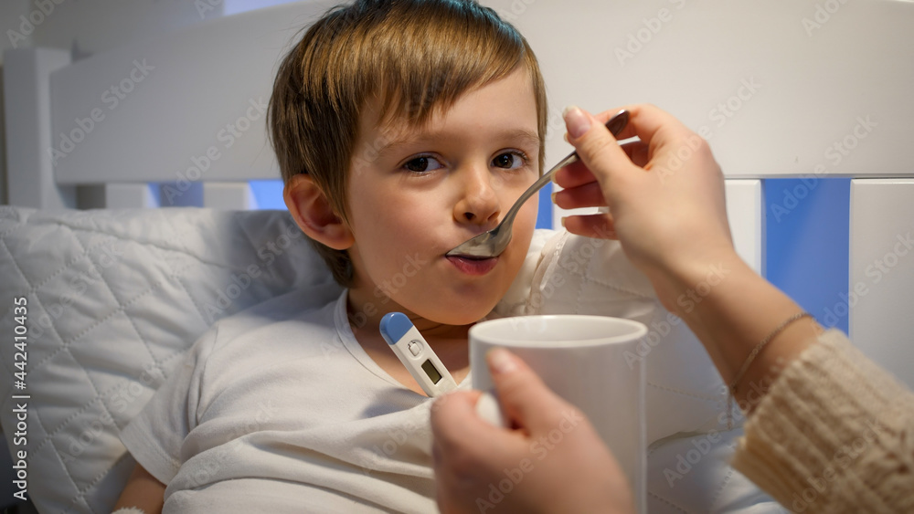Portrait of sick little boy measuring body temperature and taking curing syrup from spoon. Concept of children illness, disease and parent care Portrait of sick little boy measuring body temperature and taking curing syrup from spoon. Concept of children illness, disease and parent care