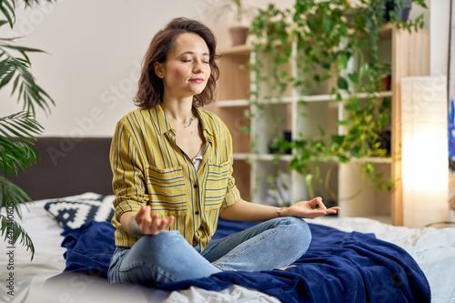 Relaxed young woman sitting on bed in Lotus pose, meditating at home