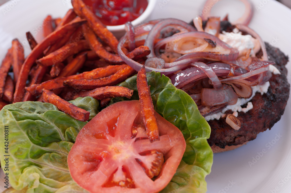 Image of a hamburger with sweet potatoes fries, tomato slice, lettuce, and onions on a white dish. Image of a hamburger with sweet potatoes fries, tomato slice, lettuce, and onions on a white dish.