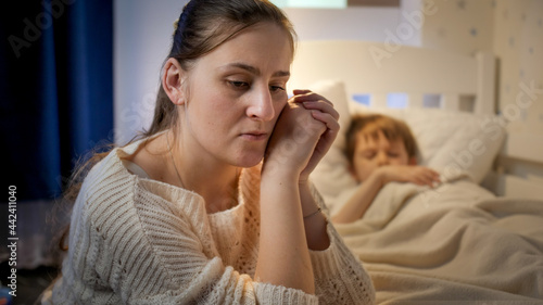 Young mother sitting next to bed of her sleeping son suffering from depression and bad thoughts. Concept of lonely mother, family problems and maternal depression