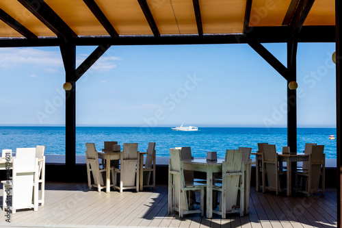 Fototapeta Naklejka Na Ścianę i Meble -  Summer resort cafe with wooden furniture with a view of the seascape with a floating motor ship. Without people