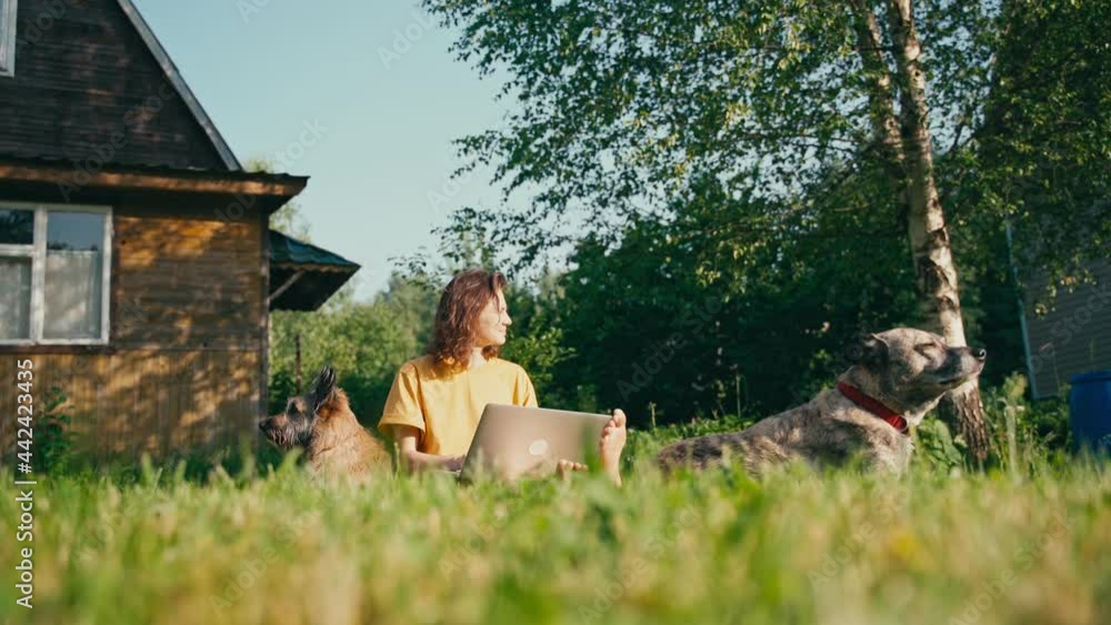 Young woman freelancer works at a laptop in the countryside while sitting on the lawn with her dogs in front of her house Young woman freelancer works at a laptop in the countryside while sitting on the lawn with her dogs in front of her house