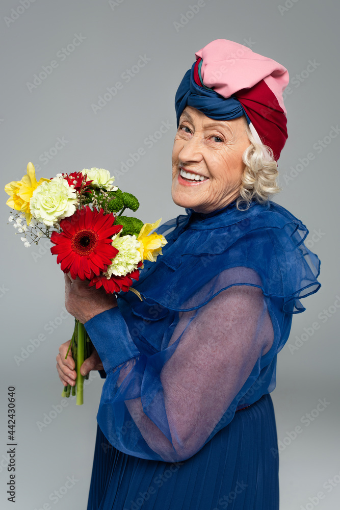 positive elderly woman in blue dress and turban holding bouquet of flowers isolated on grey positive elderly woman in blue dress and turban holding bouquet of flowers isolated on grey