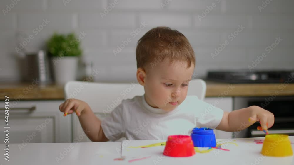 Baby boy sitting behind table and drawing with fingers covered in colorufl paint at home kitchen Baby boy sitting behind table and drawing with fingers covered in colorufl paint at home kitchen