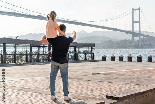 Little cute girl seat on father's shoulders in front of Bosphorus bridge (aka: 15 July Martyrs Bridge Turkish: 15 Temmuz Sehitler Koprusu) in Ortakoy, Istanbul, Turkey. Man with daughter.