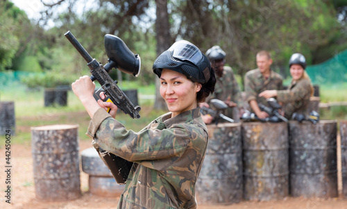Smiling asian woman paintball player in camouflage posing with gun outdoors