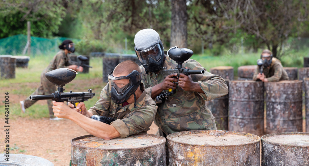 Two paintball player in camouflage and mask aiming with gun in shootout outdoors