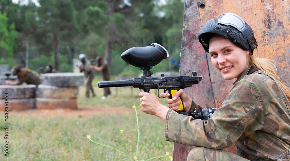 Fototapeta premium Young smiling woman paintball player in camouflage posing with gun outdoors