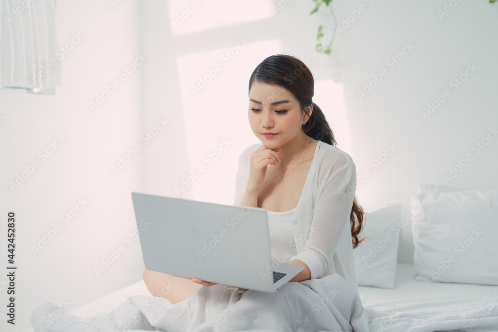 Millennial girl sitting on bed, working on laptop Millennial girl sitting on bed, working on laptop