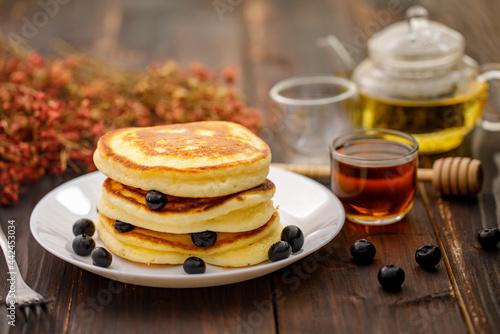 Sweet food. Stack of delicious pancakes with fork rests blueberries and honey, teapot in white plate on blur wooden background.