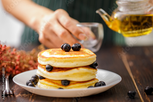 Sweet food. Stack of delicious pancakes with fork rests blueberries in white plate on blur wooden background. A holding hand teapot on the glass.