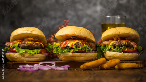Three hamburgers with beef burgers, fresh onions, fried onions.spinach, ketchup and cheese served on dark wooden chopping board  and dry leaves beer loft background.